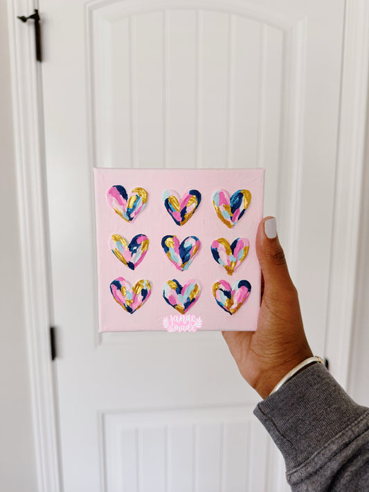 A hand holds a small square pink canvas featuring nine colorful painted hearts, with a white paneled door in the background.