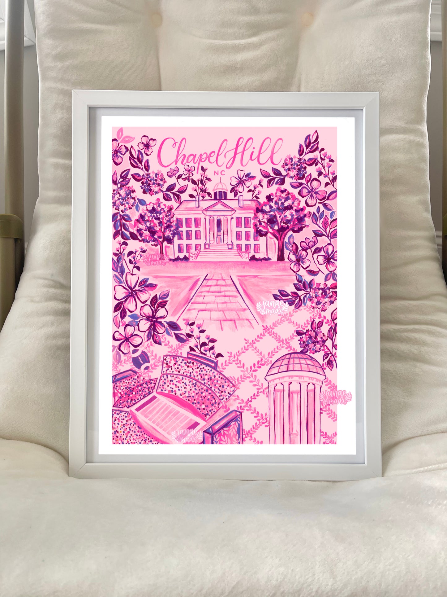 this is a vibrant pink and fuschia floral art print based on a hand painted print. The print is filled with details uniique to chapel hill north carolina. The painting is placed on a soft lounge chair and framed with a white frame.