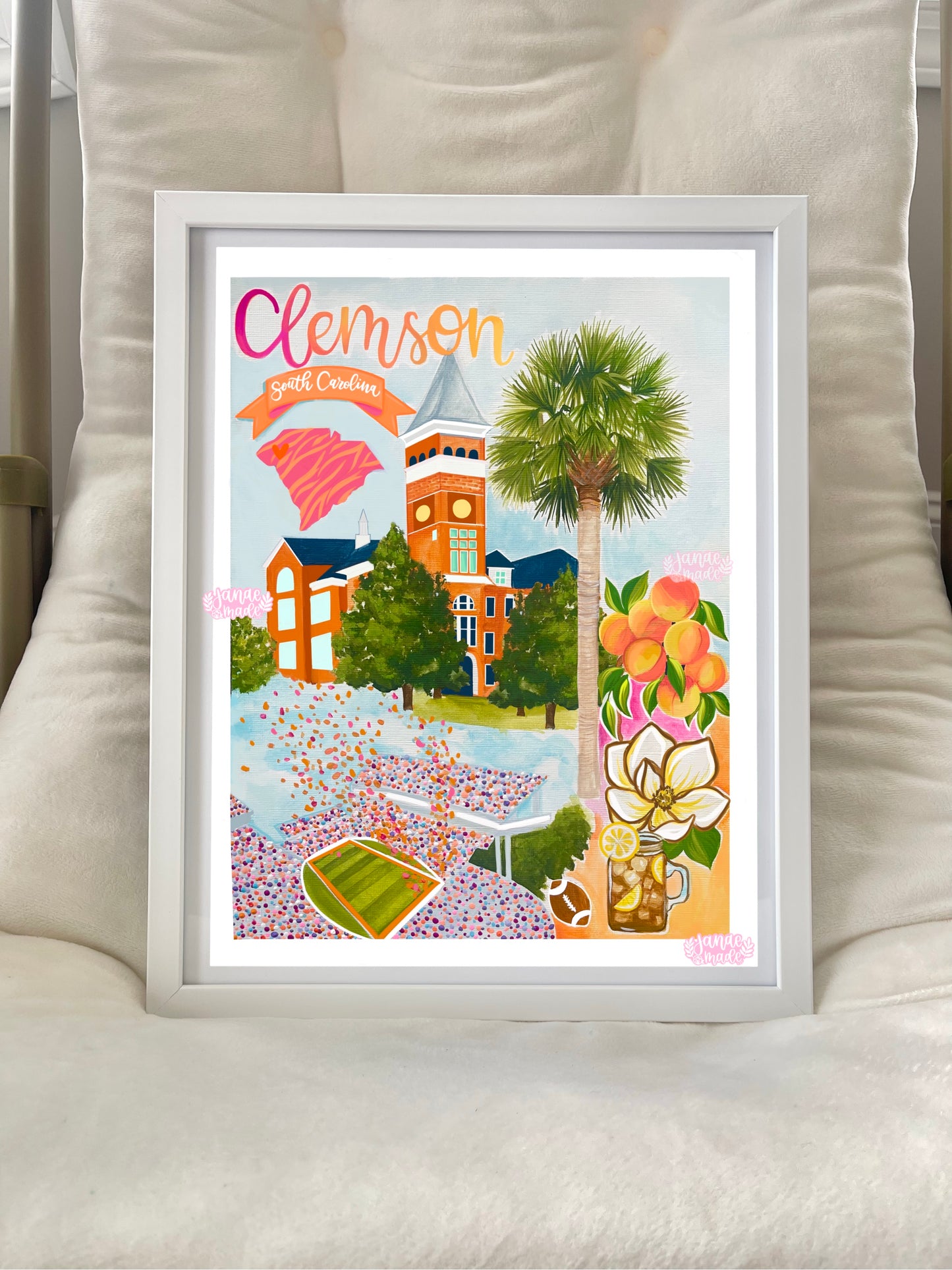 A colorful art print rests in a white frame featuring details unique to Clemson South Carolina. There is a palmetto, magnolia, a jar or sweet tea, and a football stadium. The framed piece is sitting on top of a comfy white lounge chair.