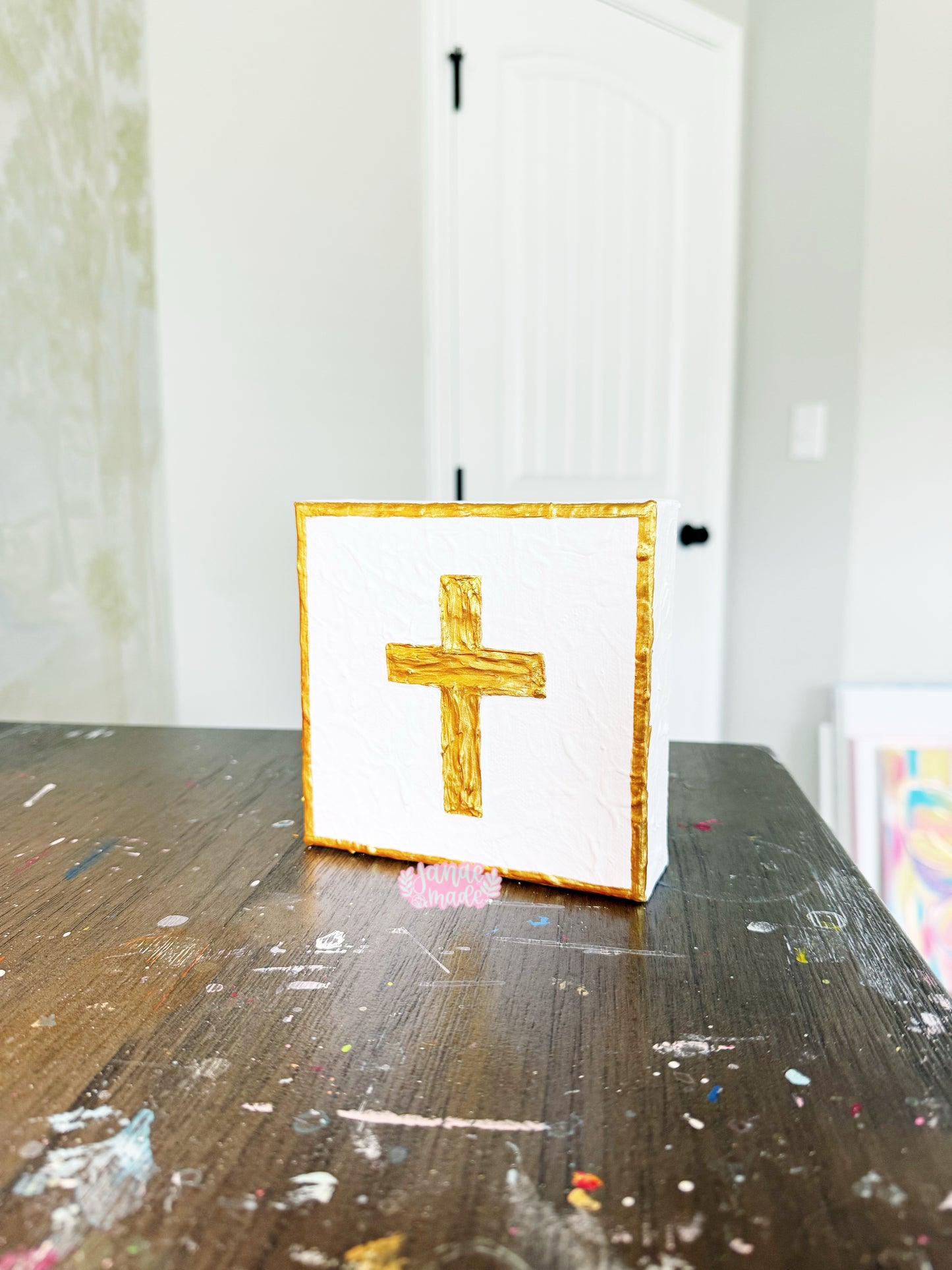 A small square white canvas featuring a textured gold cross, standing on a dark wooden table with paint splatters. A white door is visible in the softly blurred background.