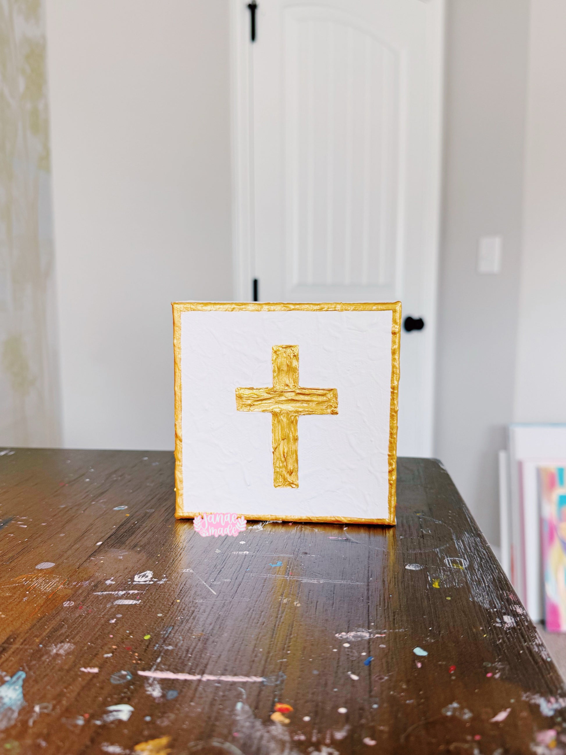 A square white canvas art print featuring a textured gold cross in the center, standing on a wooden table with paint splatters. The background shows a white paneled door and a neutral wall.