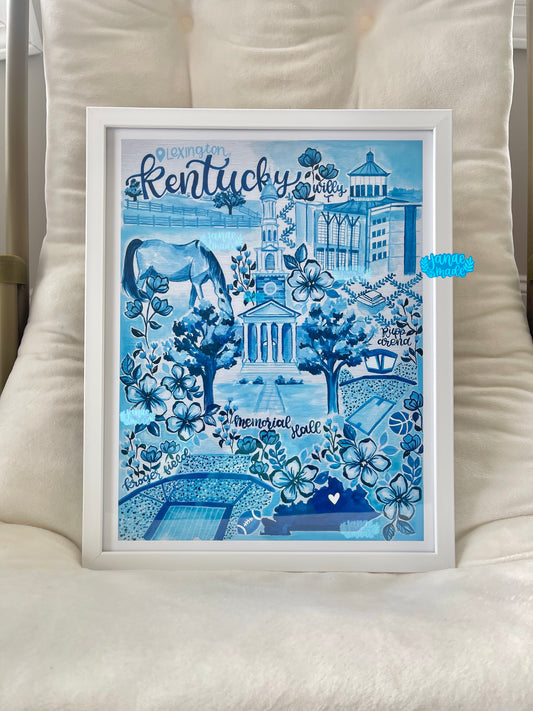 The Lexington, KY toile art print sits in a white frame on top of a soft white lounge chair. The artwork is blue filled with floral details, a bright blue state of Kentucky outline, and a watercolor style horse.