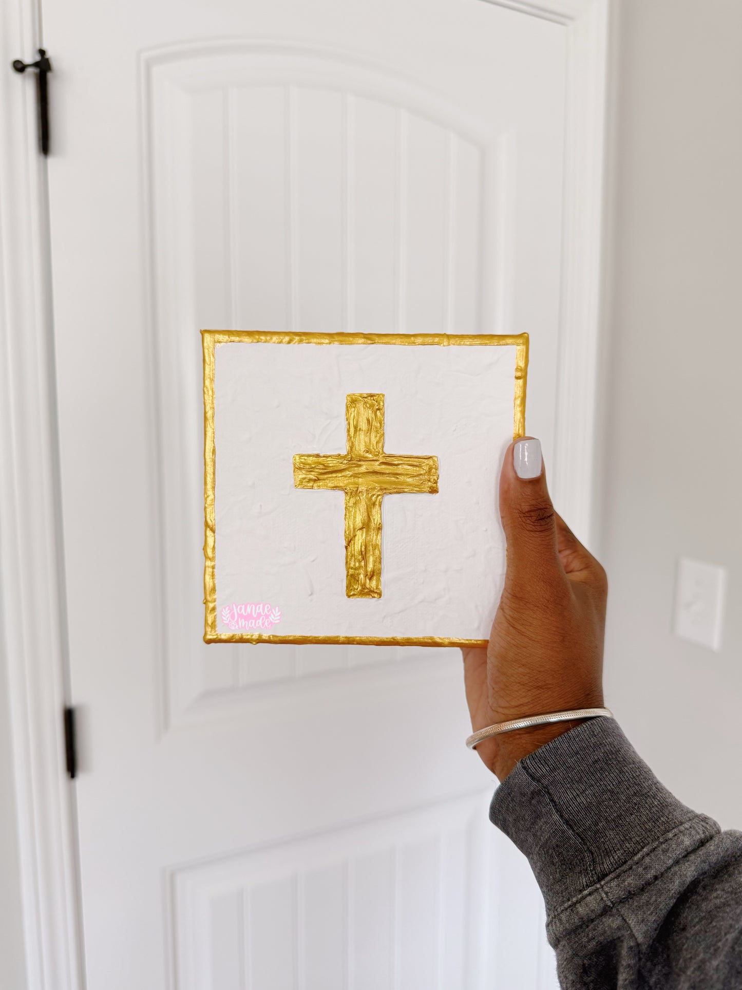 A hand holding a square white mini canvas with a textured gold cross in the center and a gold border, shown in front of a white paneled door.