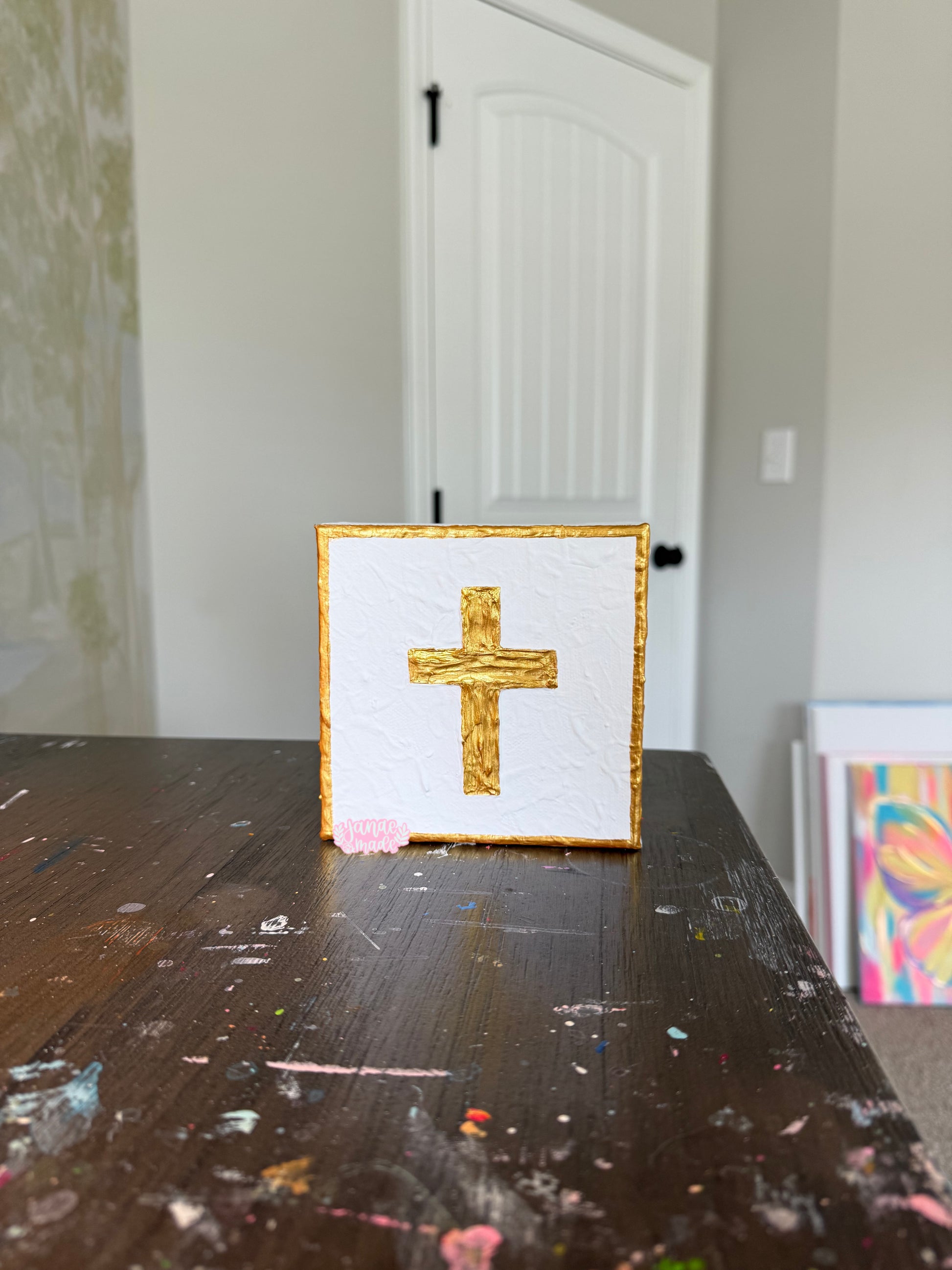 A square white canvas art print featuring a textured gold cross in the center, framed with a gold border, displayed on a dark wooden table in a room with a white door in the background.
