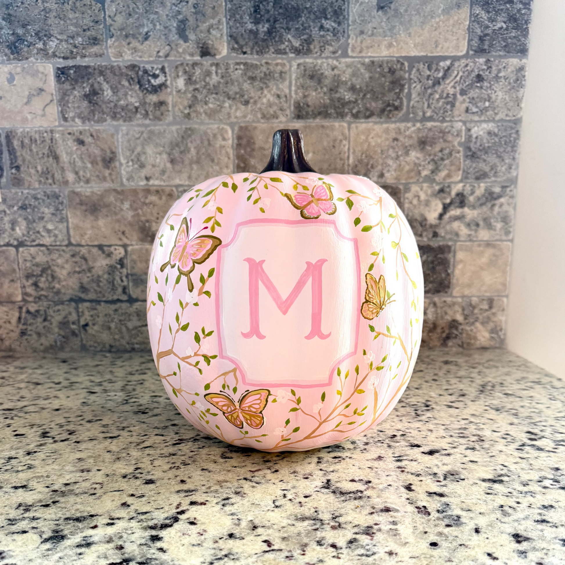 A decorative pink pumpkin with a monogrammed letter 'M' in the center, decorated with butterflies and floral patterns, sitting on a granite countertop in front of a tiled backsplash.