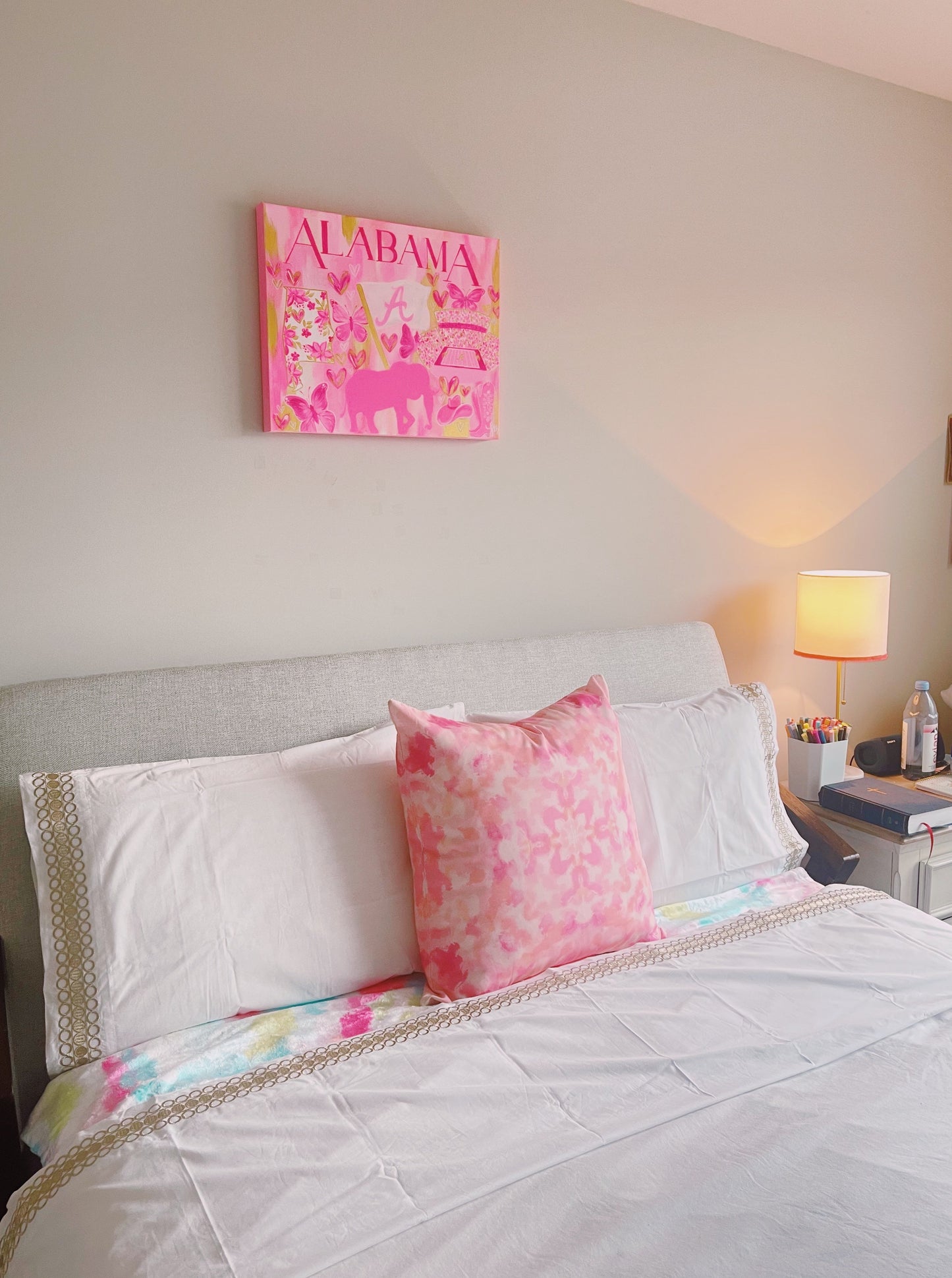 This is a pink and gold watercolor style painting with "ALABAMA" written on top in hot pink caps. There are pink butterflies, a bight pink flag with an "A" and the state of Alabama filled with floral pink and gold detailing. Under the flag is a pink elephant silhouette and there are a pair of pink and white cowgirl boots next to it. The painting sits on the wall above a bed.