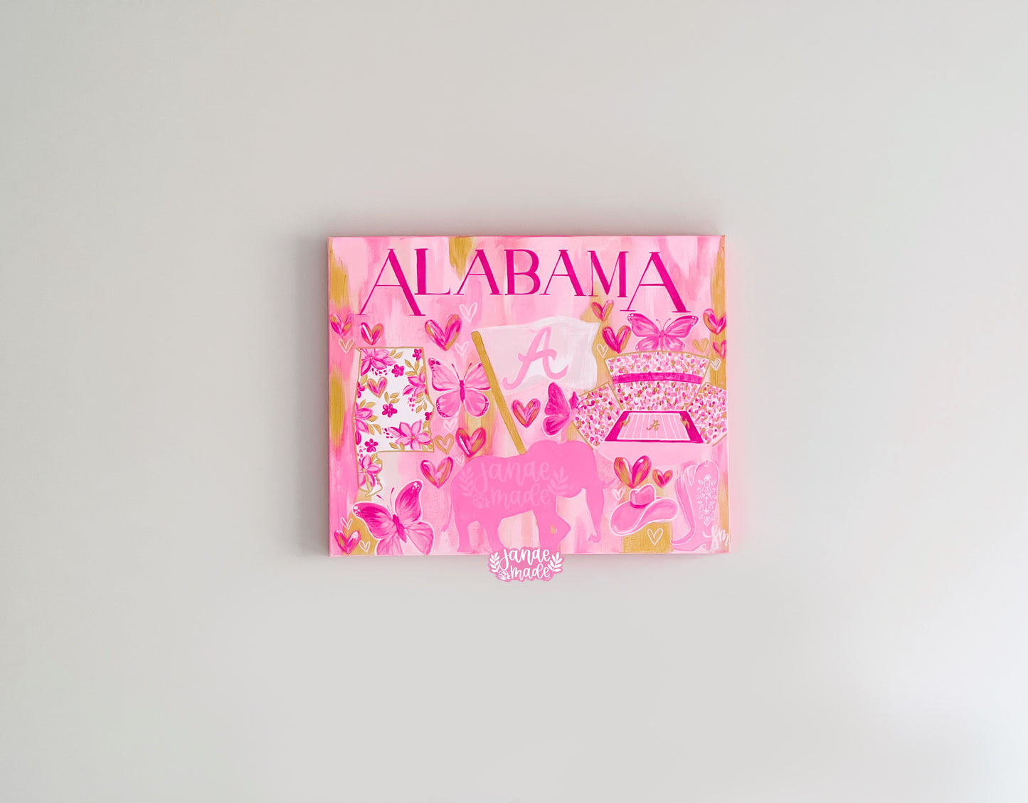 This is a pink and gold watercolor style painting with "ALABAMA" written on top in hot pink caps. There are pink butterflies, a bight pink flag with an "A" and the state of Alabama filled with floral pink and gold detailing. Under the flag is a pink elephant silhouette and there are a pair of pink and white cowgirl boots next to it.